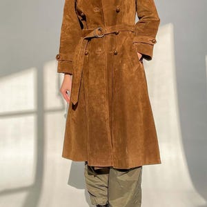 May include: A brown suede trench coat with a double-breasted design, a belt, and button details. The coat has a classic trench coat silhouette with a wide collar and long sleeves. The coat is worn with green pants and black boots.