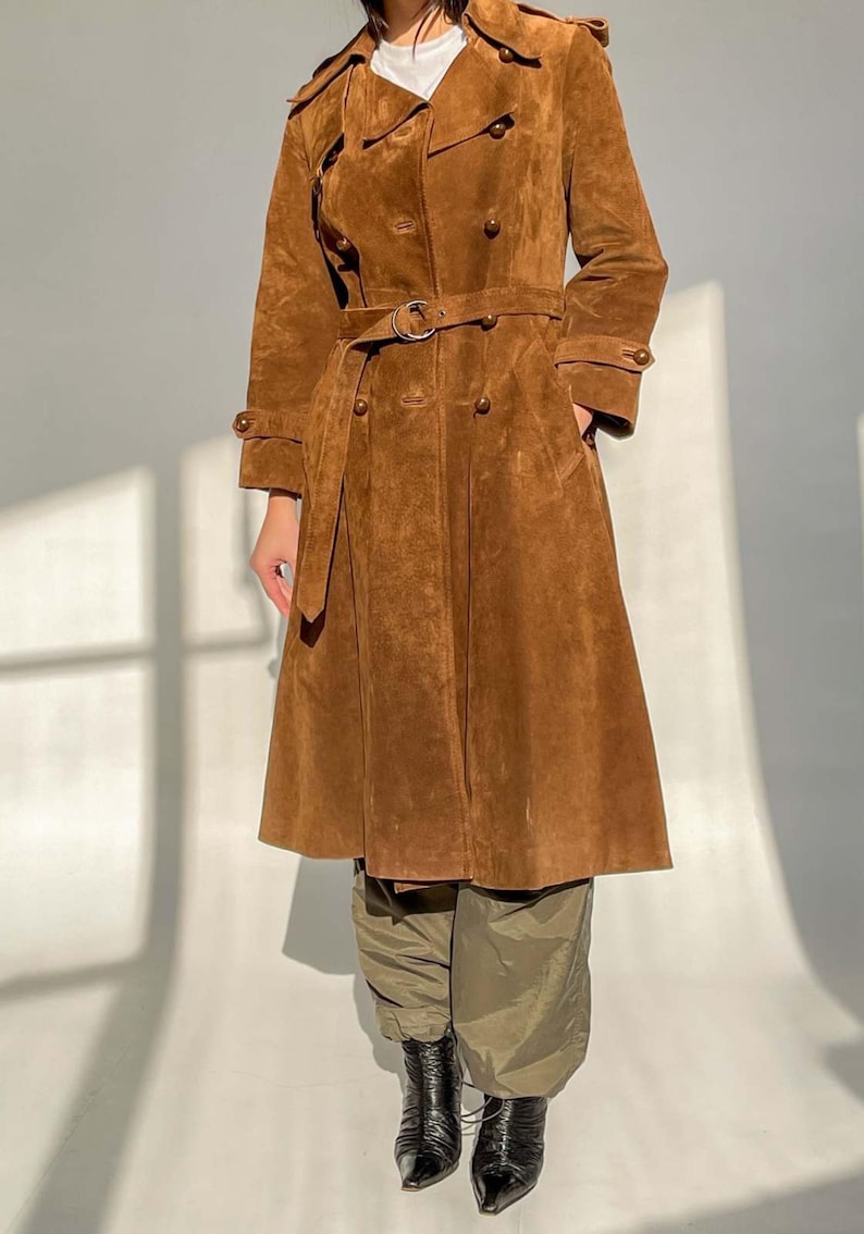 May include: A brown suede trench coat with a double-breasted design, a belt, and button details. The coat has a classic trench coat silhouette with a wide collar and long sleeves. The coat is worn with green pants and black boots.