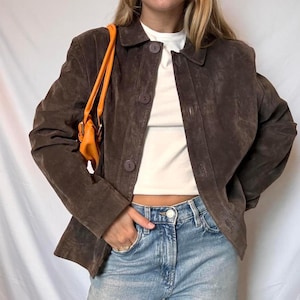 May include: A brown suede jacket with a button-up front and a collared neckline. The jacket is paired with light blue jeans and a small orange purse. The jacket has a classic, tailored fit.