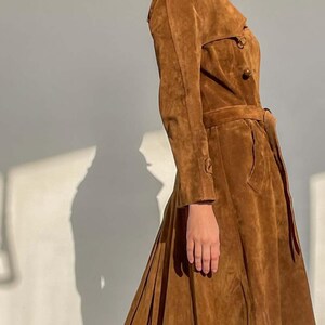 May include: A brown suede trench coat with a belted waist and button details. The coat has long sleeves and a flared skirt, creating a classic silhouette. The coat is a warm, earthy tone, perfect for a vintage or bohemian aesthetic.