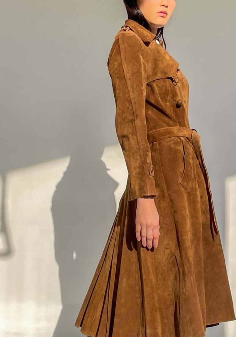 May include: A brown suede trench coat with a belted waist and button details. The coat has long sleeves and a flared skirt, creating a classic silhouette. The coat is a warm, earthy tone, perfect for a vintage or bohemian aesthetic.