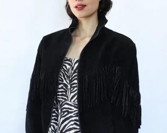 Puede incluir: Chaqueta negra de ante con flecos en las mangas. La chaqueta tiene cremallera frontal y cuello alto. La modelo lleva un vestido con estampado de cebra. Gafas de sol en la cabeza.
