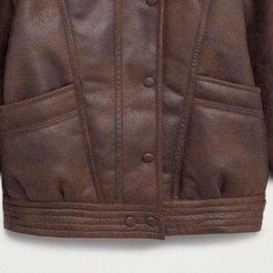 May include: A brown leather jacket with a button-up front, two front pockets, and a ribbed hem. The jacket has a classic design with a vintage look. The leather appears to be distressed, giving it a worn appearance.