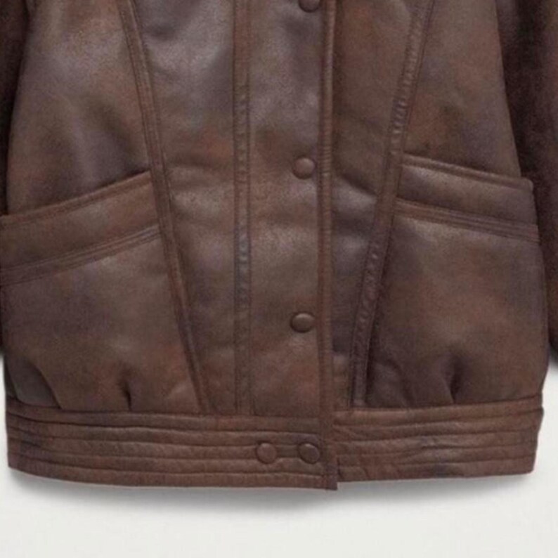 May include: A brown leather jacket with a button-up front, two front pockets, and a ribbed hem. The jacket has a classic design with a vintage look. The leather appears to be distressed, giving it a worn appearance.