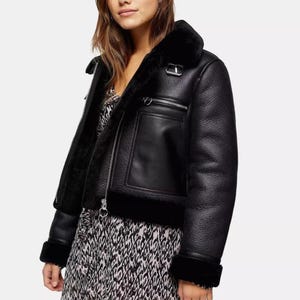 May include: Black faux leather jacket with a shearling lining and collar. The jacket features a zip closure, a pocket on the chest, and a buckle detail on the collar. The cuffs and hem are trimmed with black shearling.