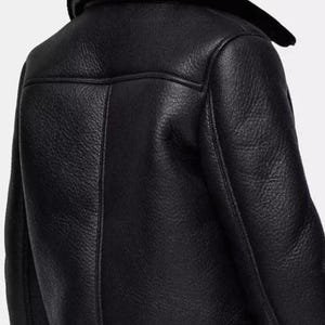 May include: Close-up of a black leather jacket with a shearling collar. The jacket has a textured surface and visible stitching details. The back of the jacket is shown, highlighting the design and construction of the garment.