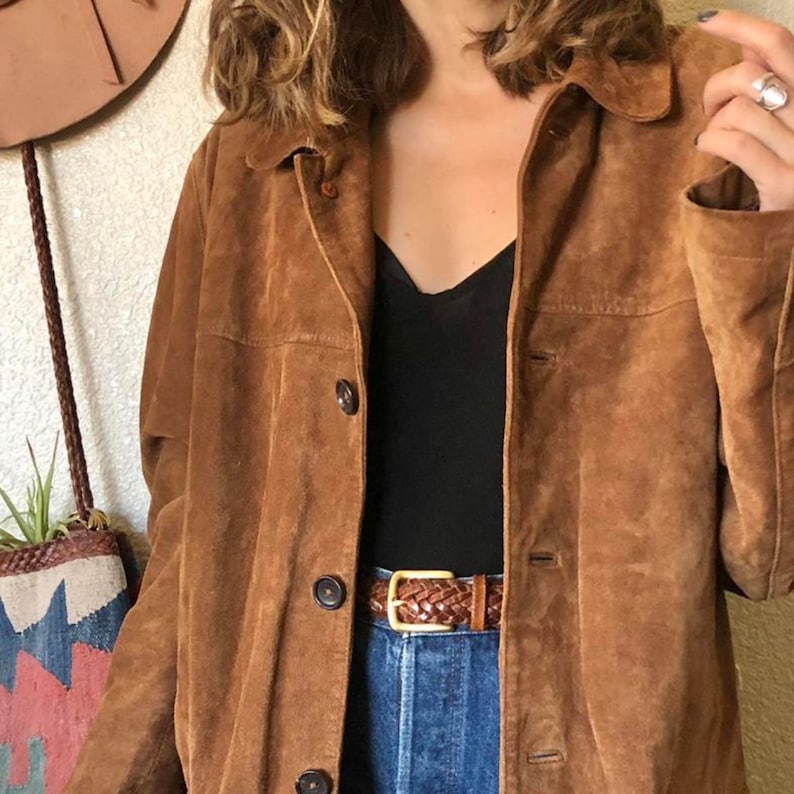 May include: A brown suede jacket with a button-up front and a collared neckline. The jacket is worn over a black top and blue jeans, with a brown woven belt. The jacket has a classic, vintage style.