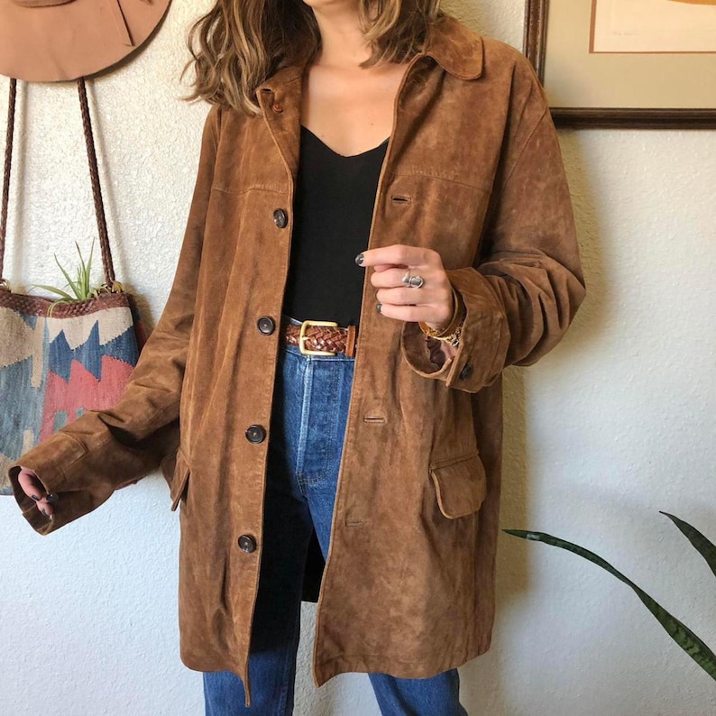 May include: A brown suede coat with a button-down front, two front pockets, and a collared neckline. The coat is worn over a black top and blue jeans. A brown leather belt with a gold buckle is visible.