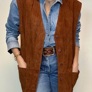 May include: A brown suede vest with a button-up front and two front pockets. The vest is worn over a denim shirt and blue jeans. A brown belt with a silver buckle is visible.