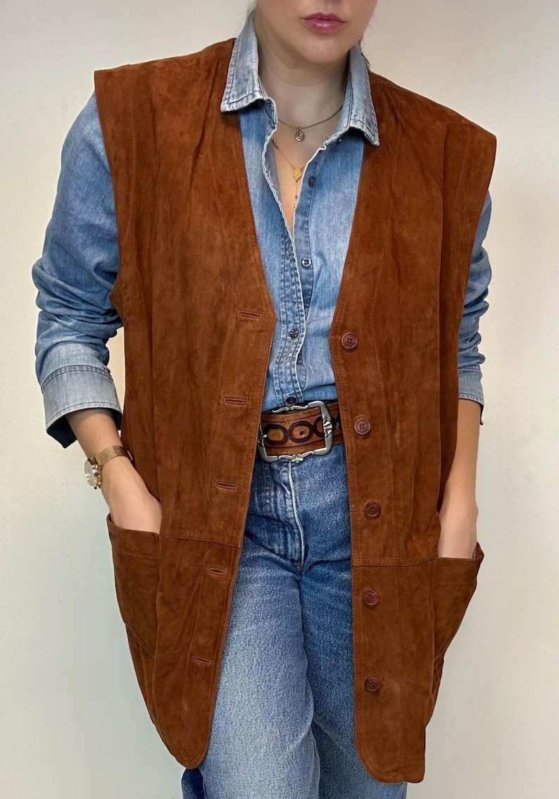 May include: A brown suede vest with a button-up front and two front pockets. The vest is worn over a denim shirt and blue jeans. A brown belt with a silver buckle is visible.