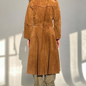 May include: A brown suede trench coat with a belted waist and pleated skirt. The coat has long sleeves and a classic collar. The model is wearing olive green pants and black high-heeled boots. The coat is a vintage style.