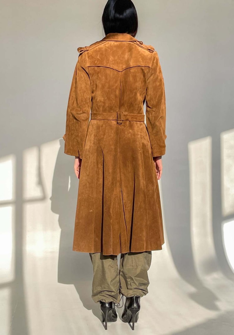 May include: A brown suede trench coat with a belted waist and pleated skirt. The coat has long sleeves and a classic collar. The model is wearing olive green pants and black high-heeled boots. The coat is a vintage style.