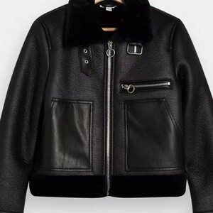 May include: Black faux leather jacket with a shearling collar and lining. The jacket features a silver zipper closure, two front pockets, and a zippered chest pocket. The jacket has two decorative buttons on the left side.