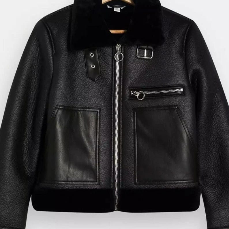 May include: Black faux leather jacket with a shearling collar and lining. The jacket features a silver zipper closure, two front pockets, and a zippered chest pocket. The jacket has two decorative buttons on the left side.