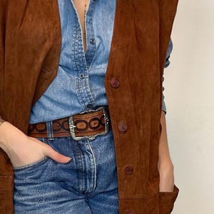 May include: A brown suede vest with button closures over a denim shirt and blue jeans. A brown leather belt with a silver buckle is worn at the waist. The outfit is accessorized with a gold necklace.