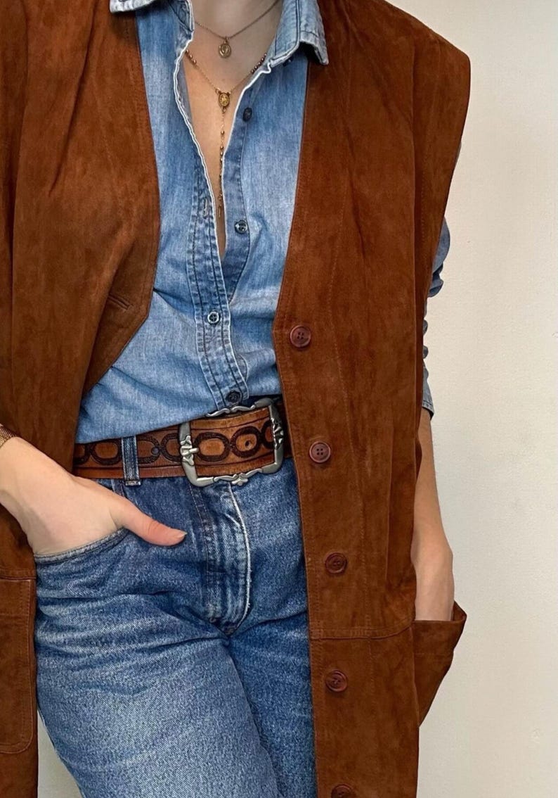 May include: A brown suede vest with button closures over a denim shirt and blue jeans. A brown leather belt with a silver buckle is worn at the waist. The outfit is accessorized with a gold necklace.