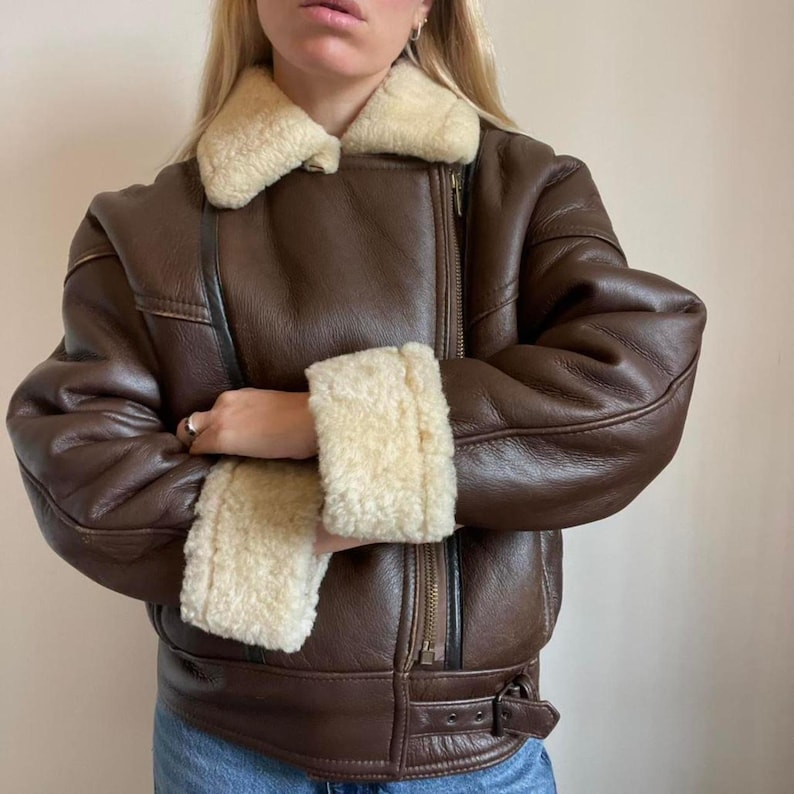 May include: A brown leather jacket with a cream-colored shearling collar and cuffs. The jacket features a zipper closure, a belt at the waist, and a classic aviator style. The jacket is a medium weight and is suitable for cooler weather.