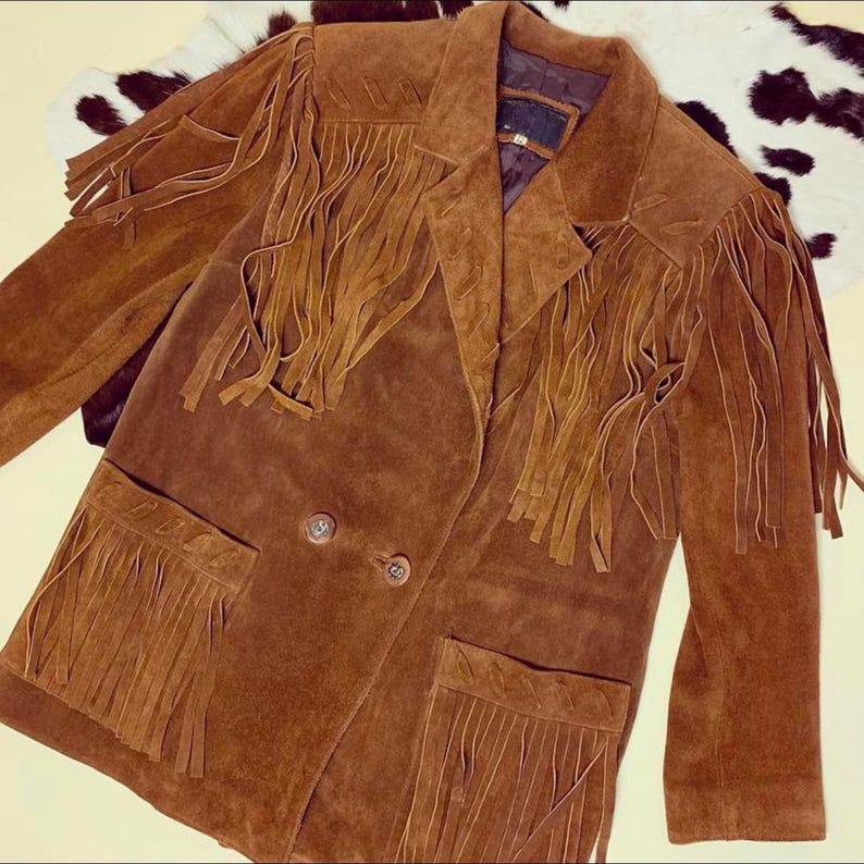 May include: A brown suede jacket with fringe detailing on the chest, pockets, and sleeves. The jacket has a double-breasted closure with two buttons and a notched collar. The jacket is a medium brown color.
