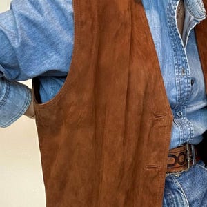 May include: A brown suede vest with a classic design. The vest has a button-up front and a tailored fit. The vest is layered over a light blue denim shirt. The vest is a warm, earthy tone, perfect for layering.