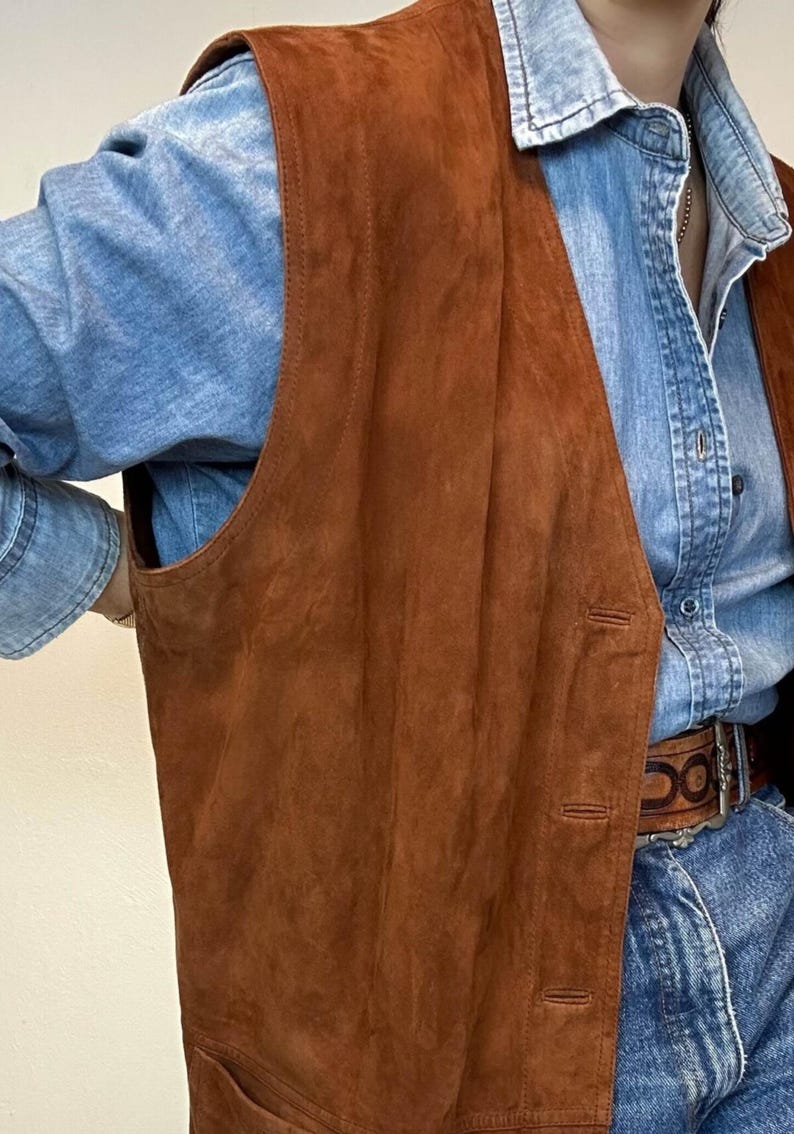 May include: A brown suede vest with a classic design. The vest has a button-up front and a tailored fit. The vest is layered over a light blue denim shirt. The vest is a warm, earthy tone, perfect for layering.