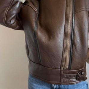May include: A brown leather jacket with a zipper closure and a buckle detail at the waist. The jacket has a shearling lining visible at the collar and cuffs. The leather appears worn, with visible stitching and panels.