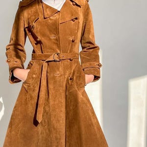 May include: A brown suede trench coat with a double-breasted design, featuring a belt at the waist and pockets. The coat has button details on the sleeves and a classic collar. The coat is knee-length.