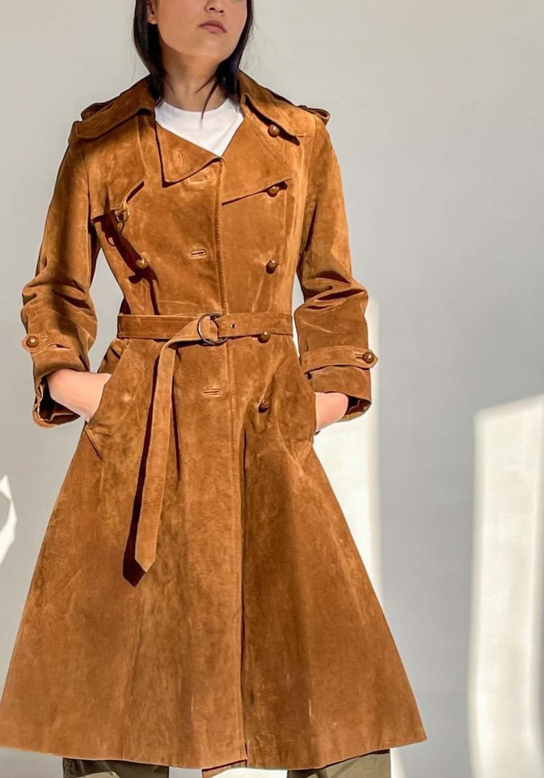 May include: A brown suede trench coat with a double-breasted design, featuring a belt at the waist and pockets. The coat has button details on the sleeves and a classic collar. The coat is knee-length.