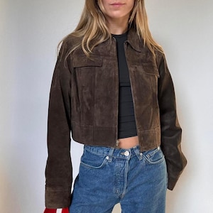 Peut inclure: Veste en daim marron avec un col et deux poches avant. La veste est courte et possède une fermeture éclair. Le modèle porte un haut noir et un jean bleu.
