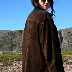 May include: A dark brown suede jacket with a classic collar and long sleeves. The jacket is worn by a person wearing sunglasses, with a mountain range and blue sky in the background.