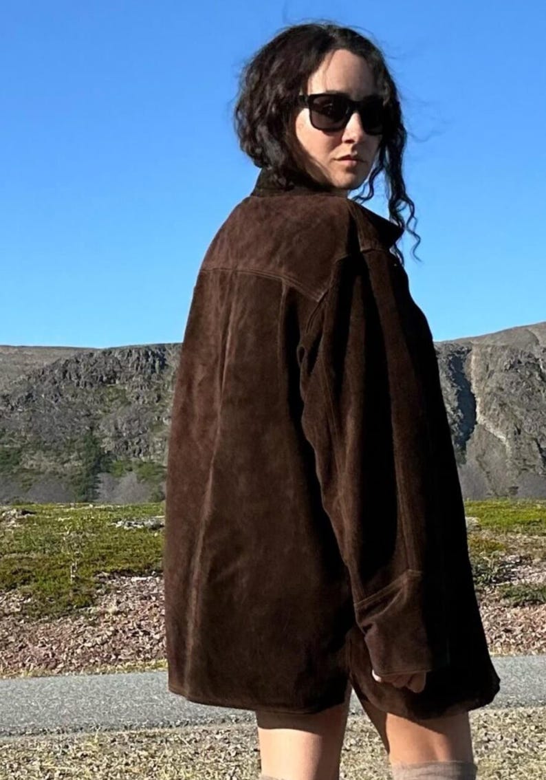 May include: A dark brown suede jacket with a classic collar and long sleeves. The jacket is worn by a person wearing sunglasses, with a mountain range and blue sky in the background.