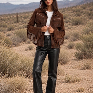 May include: A brown suede jacket with long fringe detailing on the chest and sleeves, featuring silver-tone metal zippers and buttons. The jacket is worn over a plain white t-shirt paired with black leather trousers and brown suede ankle boots.