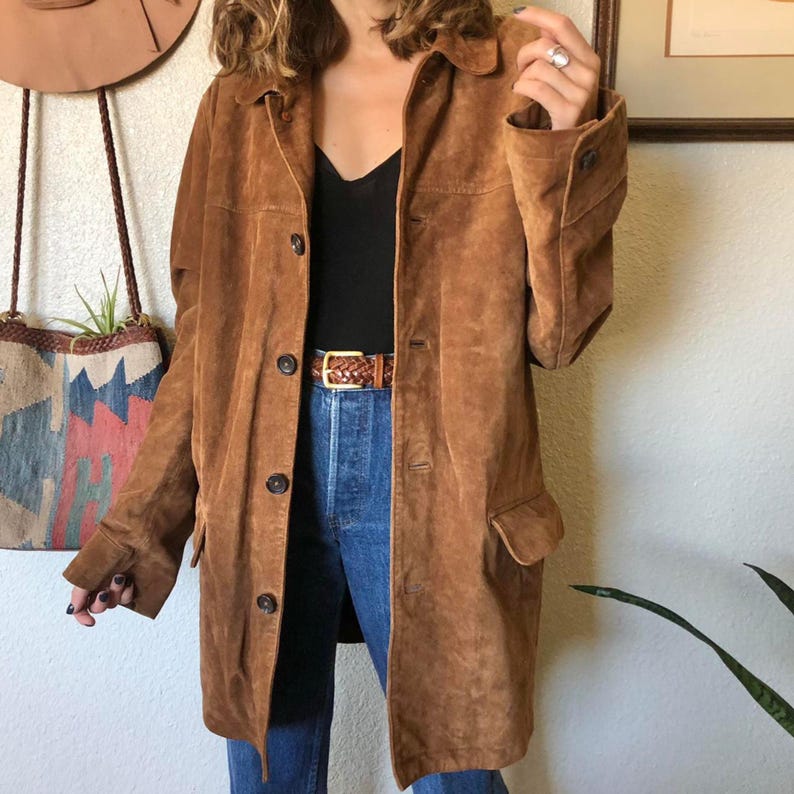 May include: A brown suede coat with a button-up front, two front pockets, and a collared neckline. The coat is knee-length and has long sleeves. A brown leather belt with a gold buckle is visible.