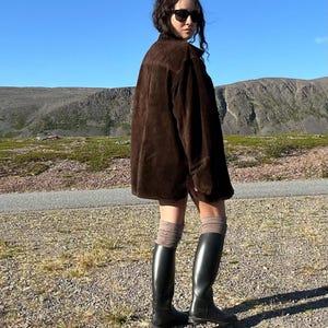 May include: A person wearing a dark brown suede jacket, black sunglasses, and black knee-high boots. The person is standing on a gravel path with a mountain range and blue sky in the background. The boots are approximately 16 inches tall.