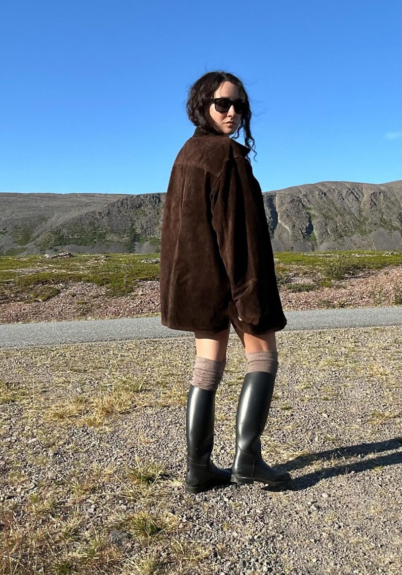May include: A person wearing a dark brown suede jacket, black sunglasses, and black knee-high boots. The person is standing on a gravel path with a mountain range and blue sky in the background. The boots are approximately 16 inches tall.