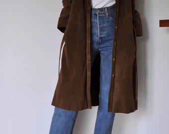 May include: A brown suede long coat with a button-down front, paired with blue jeans and white sneakers. The coat has a collared neckline and long sleeves. The jeans are a classic denim wash.