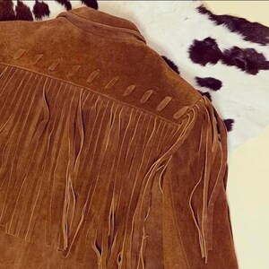 May include: A brown suede jacket with fringe detailing across the back and sleeves. The jacket has a classic western style, with a collar and a button-up front. The fringe adds a decorative element to the jacket. The jacket is displayed on a white and black cowhide.