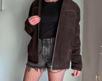 Peut inclure: Veste en daim marron avec fermeture éclair, deux poches poitrine et deux poches latérales. La veste est portée sur un haut noir et un short en jean. La tenue est complétée par des bas résille.