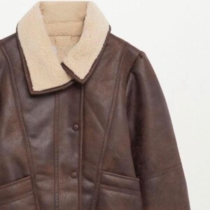 May include: A brown faux leather jacket with a cream-colored shearling collar. The jacket has a button-up front and a tailored fit. The shearling collar provides a warm and cozy look. The jacket is a classic outerwear piece.