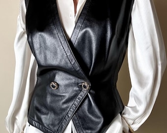 May include: Black leather vest with gold-tone buttons, worn over a white collared shirt. The vest has a double-breasted design and a tailored fit. A gold chain necklace and bracelet complete the look. The outfit is a classic, stylish ensemble.