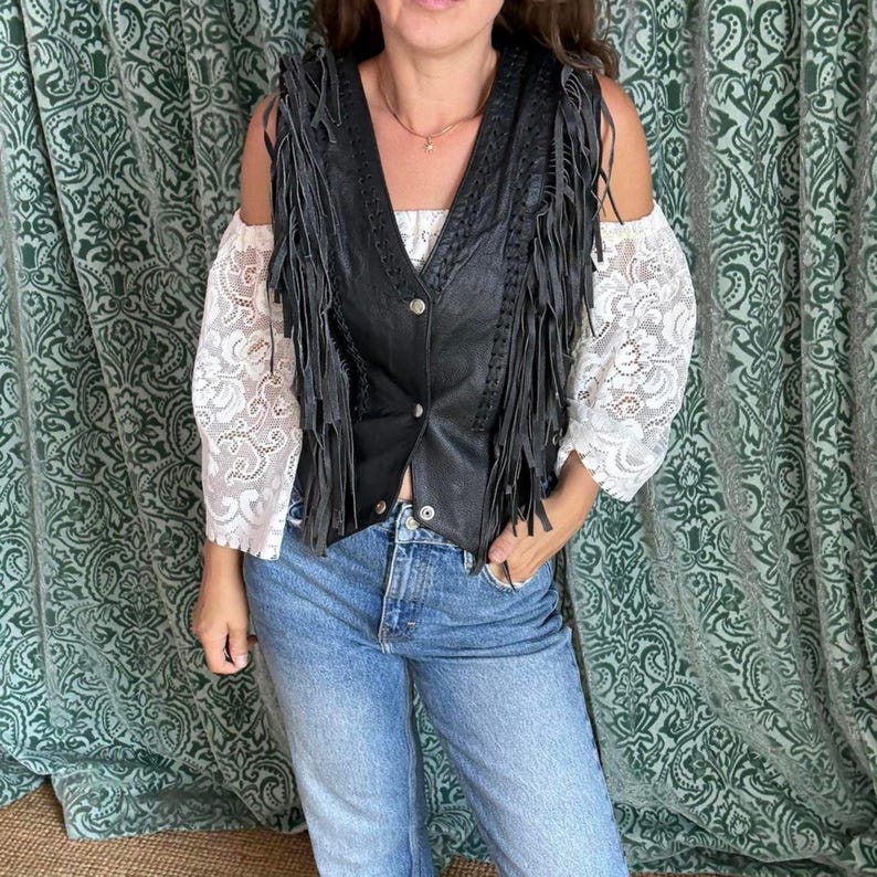 May include: Black leather vest with fringe detail, V-neck and button closure. Worn over a white lace top and blue jeans. The style evokes a bohemian or Western aesthetic. The vest's fringe adds a distinctive texture and movement.
