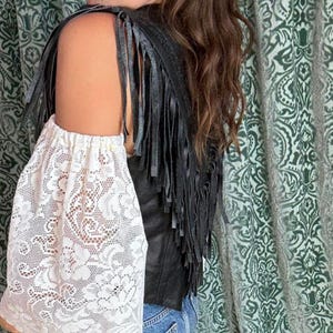 May include: A black leather vest with fringe detail and a white lace sleeve. The sleeve is made of a delicate floral lace pattern. The vest is paired with blue jeans.
