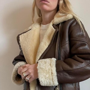 May include: A brown leather jacket with a cream-colored shearling lining and trim. The jacket features a zipper closure and a classic aviator style. The cuffs and collar are lined with the same cream-colored shearling.
