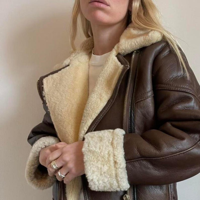 May include: A brown leather jacket with a cream-colored shearling lining and trim. The jacket features a zipper closure and a classic aviator style. The cuffs and collar are lined with the same cream-colored shearling.