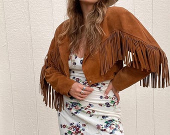 Pode incluir: Casaco de camurça castanho com franjas e chapéu de abas largas. O casaco é curto e tem franjas compridas nas mangas. Um vestido com estampa floral é usado por baixo.