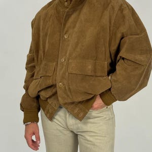 May include: A brown suede bomber jacket with a button-up front, two front pockets, and a ribbed collar and cuffs. The jacket is worn with light-colored pants. The overall style is vintage.