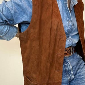May include: A long, brown suede vest with a button-up front and two front pockets. The vest is worn over a denim shirt and jeans. The vest appears to be vintage and has a classic, timeless style.