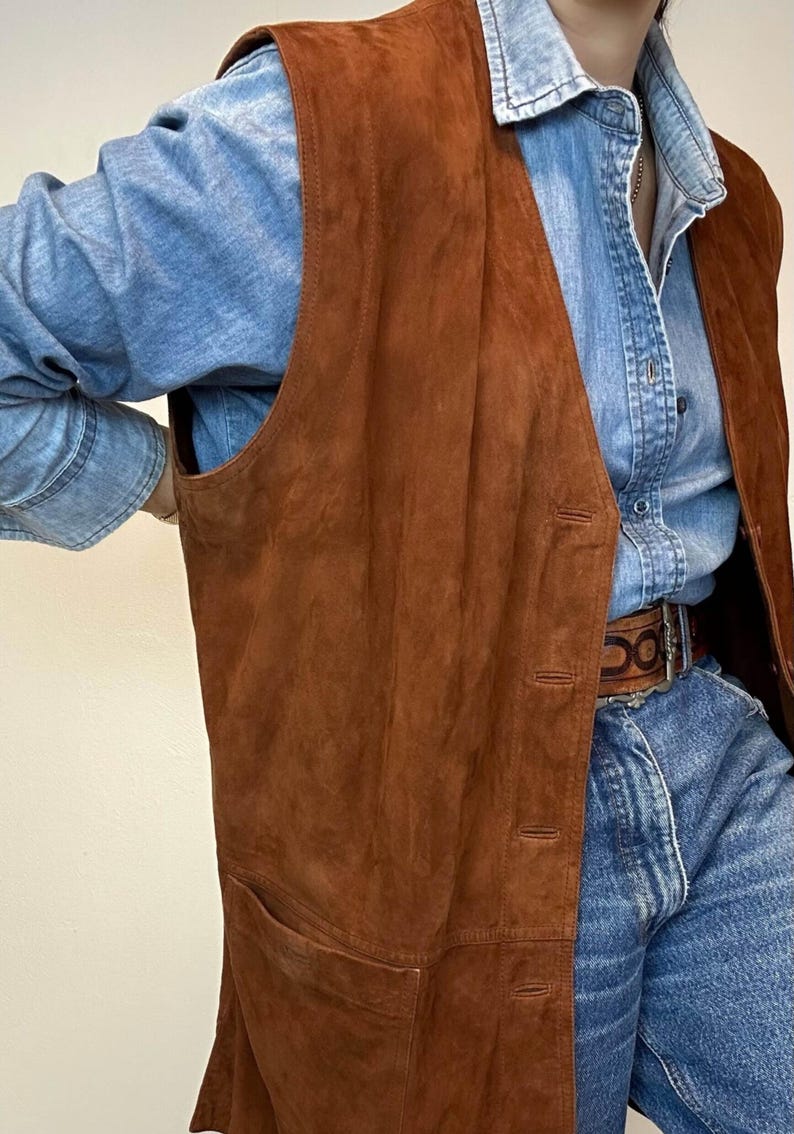 May include: A long, brown suede vest with a button-up front and two front pockets. The vest is worn over a denim shirt and jeans. The vest appears to be vintage and has a classic, timeless style.