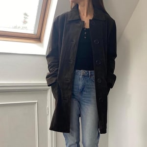 May include: A dark brown suede coat with a button-up front and a collared neckline. The coat is knee-length and has pockets. The person is wearing a black top and blue jeans. The coat is a classic outerwear piece.