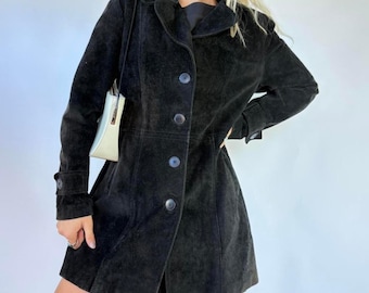 May include: A black suede coat with a button-up front, long sleeves, and a notched collar. The coat is knee-length and has a fitted silhouette. A small, off-white purse hangs from the shoulder.