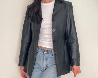 Könnte beinhalten: Ein schwarzer Lederblazer mit klassischem Design. Der Blazer hat einen Reverskragen, drei Knöpfe und lange Ärmel. Das Model trägt ein weißes Oberteil und blaue Jeans.